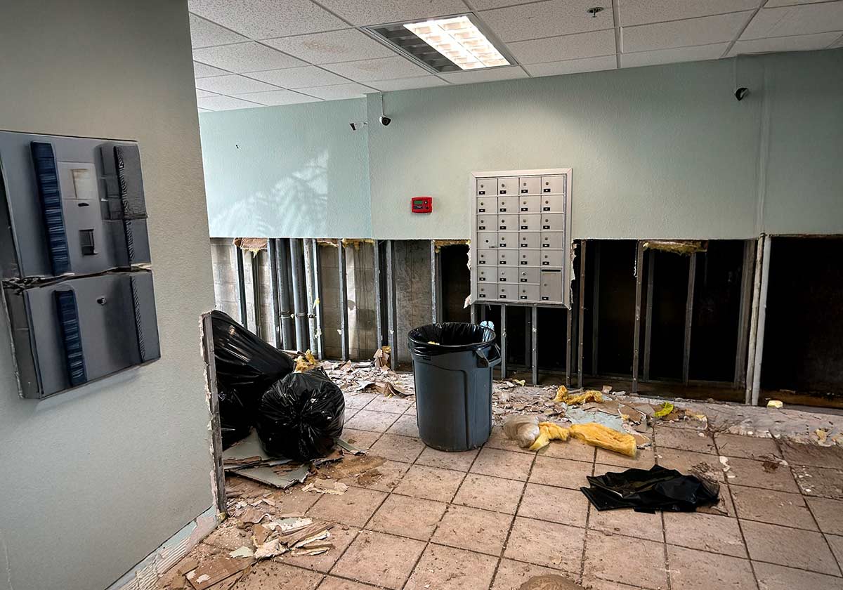 A Damaged Room With Missing Drywall, Exposed Insulation, And Debris On The Tile Floor. A Few Trash Bags And A Bin Are Present. A Panel Of Small Metal Mailboxes Is Mounted On The Wall. A Fluorescent Light Is On The Ceiling.