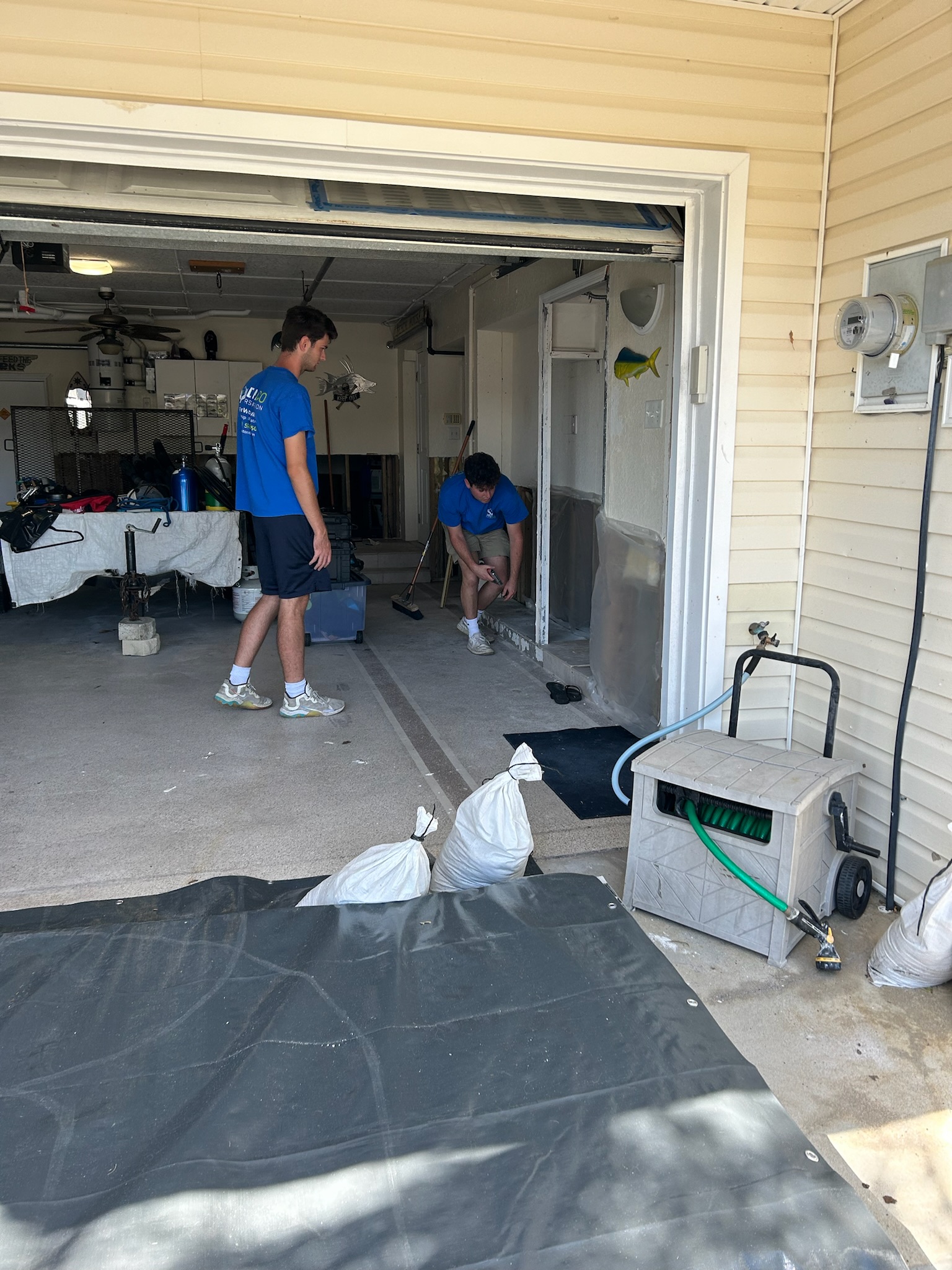 Two Restoration Workers Repairing A Residential Garage Interior With Tools And Cleanup Bags Visible. Two Restoration Workers Repairing A Residential Garage Interior With Tools And Cleanup Bags Visible.