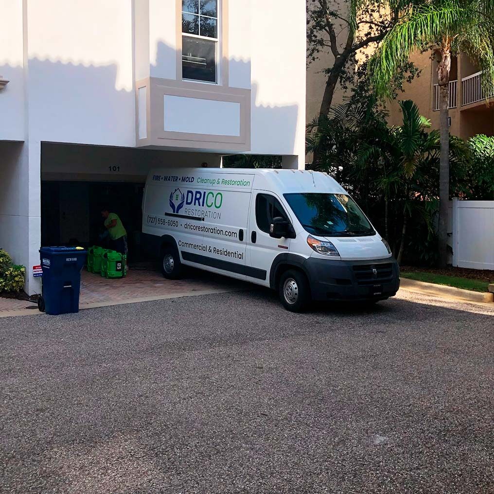 A White Van With Drico Restoration And Contact Details Parked Halfway Into A Residential Garage. A Person Is Visible Doing Work Inside The Garage. Nearby Are Recycling And Trash Bins. The Surrounding Area Has Trees And Apartment Buildings.