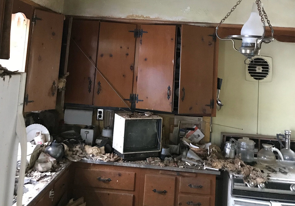 A Cluttered, Neglected Kitchen With A Vintage Tv, Old Appliances, And Debris Covering The Countertop. The Wooden Cabinets Are Worn And The Walls Appear Stained. A Broken Hanging Light Fixture And A Vintage Fan Are Visible.