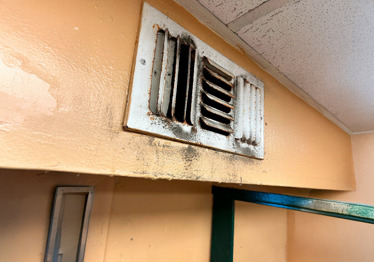 A Dirty Air Vent On A Beige Wall Shows Signs Of Neglect, With Dust And Grime Buildup. The Ceiling Is Made Of Drop Tiles. A Small Portion Of A Green-Framed Glass Structure Is Visible In The Lower Part Of The Image.