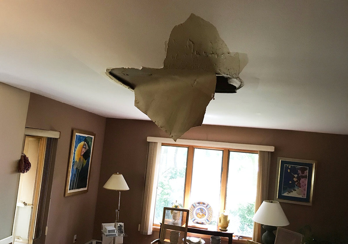 A Living Room With A Damaged Ceiling, Where A Large Piece Of Plaster Has Fallen, Revealing A Dark Hole. The Room Has Beige Walls, A Window With Curtains, Various Artworks, Two Lamps, And Several Decorative Items.