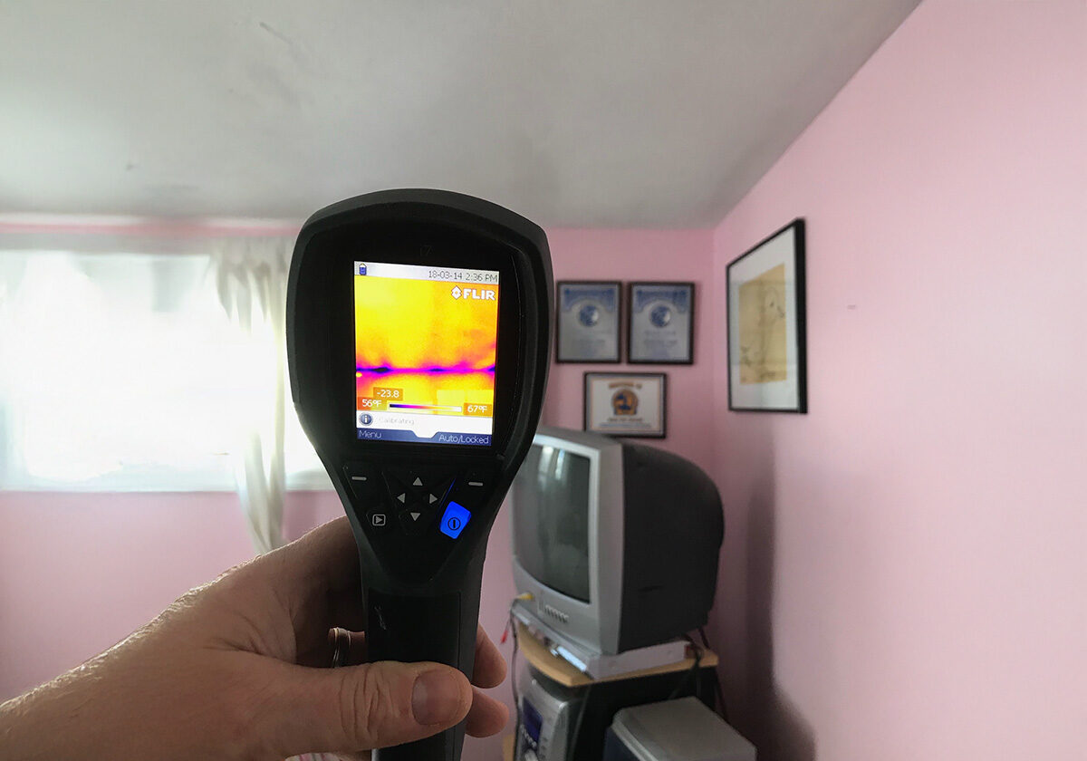 Water,-Fire,-And-Mold-Remediation-Service-Areas2 A Person Holds A Thermal Imaging Camera In A Pink Room, Focusing On A Wall. The Camera Display Shows A Thermal Gradient. A Small Tv And Framed Pictures Are Visible On A Shelf And Wall In The Background.