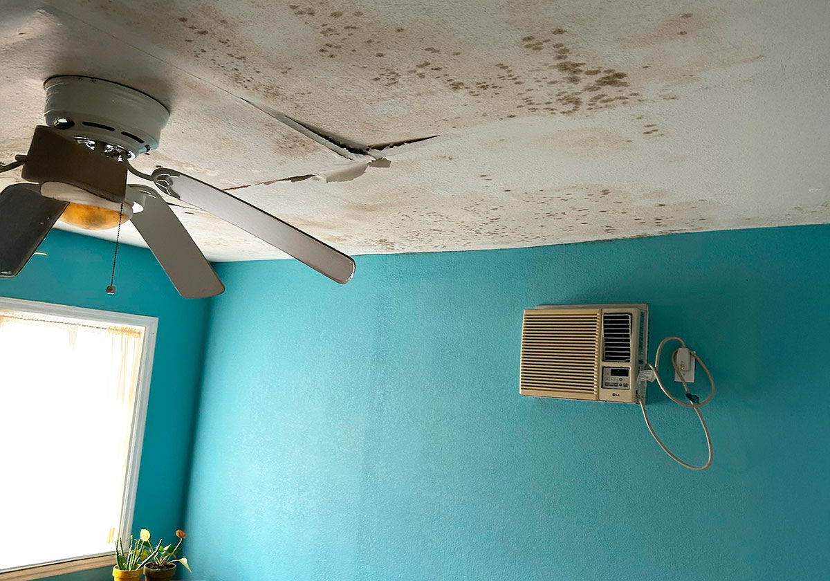 Interior Of A Room Featuring A Ceiling Fan And Cracked, Stained Ceiling, With A Wall-Mounted Air Conditioner Against A Turquoise Wall.