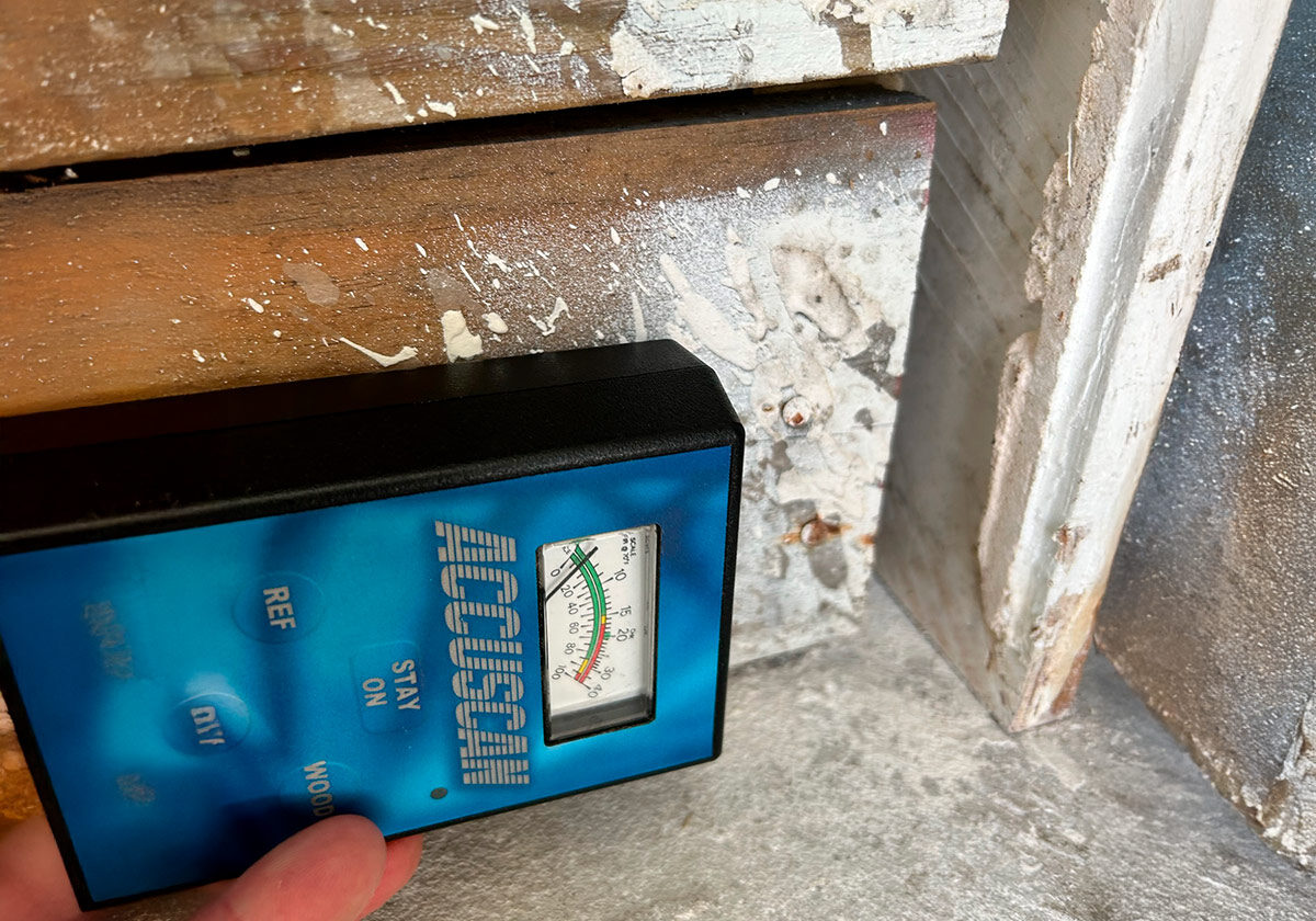 A Hand Holds A Blue Moisture Meter Against Weathered Wooden Surfaces, Measuring Moisture Levels With An Analog Gauge.