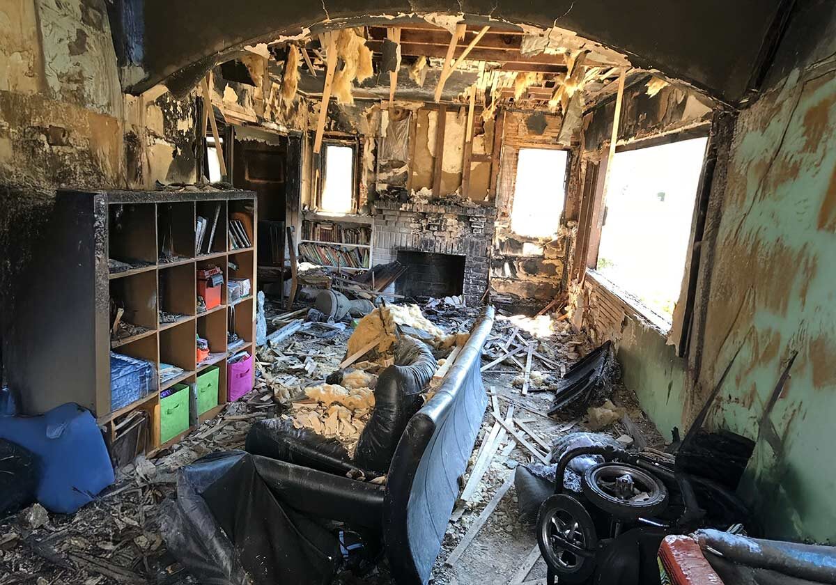 A Room Severely Damaged By Fire, Showing Charred Walls And Ceiling. Debris And Insulation Litter The Floor. A Burnt Sofa Faces A Fireplace. Bookshelves Hold Some Items, And A Childs Toy Car Is Partially Visible Among The Wreckage.