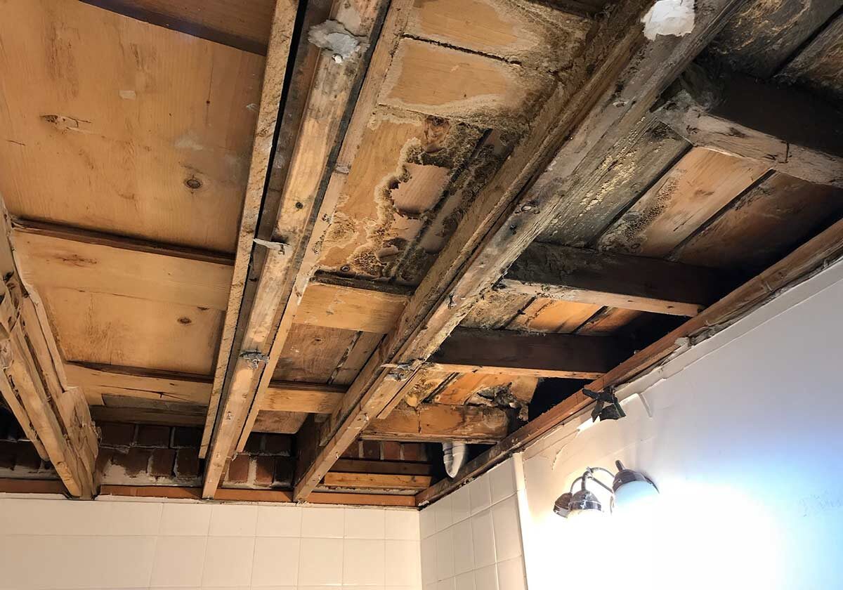 Water,-Fire,-And-Mold-Remediation-Service-Areas4 Exposed Wooden Beams And Ceiling Joists Showing Signs Of Water Damage And Decay, With Missing Ceiling Panels And Visible Light Fixtures. The Walls Are Tiled, Revealing Some Brickwork At The Upper Section.