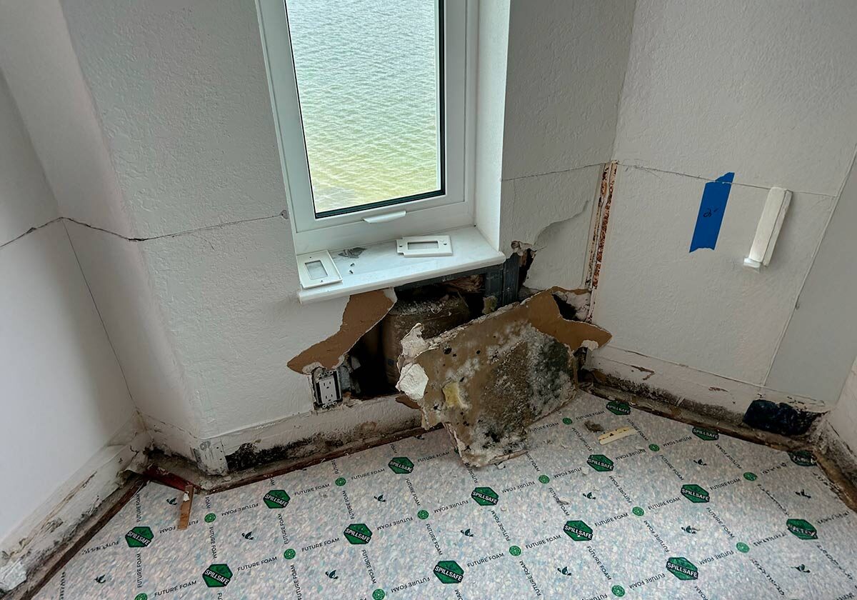 Water,-Fire,-And-Mold-Remediation-Service-Areas6 Room With A Damaged Wall And Exposed Insulation Below A Window. Floor Is Covered With Protective Plastic Sheets. Wall Has A Partial Hole With Some Mold, And Theres Blue Tape On The Right.