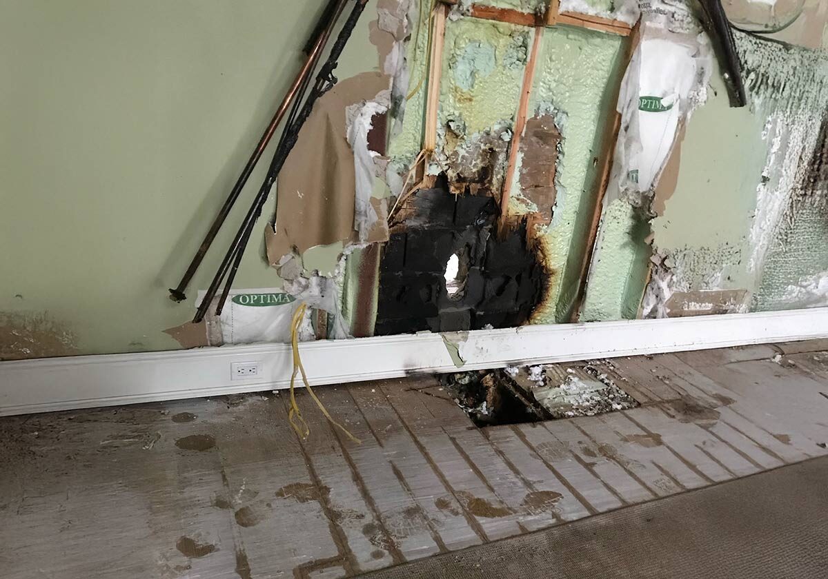 Damaged Interior Wall With A Large Hole Exposing Insulation And Wooden Beams. Water Stains And Mold Are Visible On The Green Wall. Loose Wires And Pipes Protrude, And The Hardwood Floor Is Partially Ripped Up.