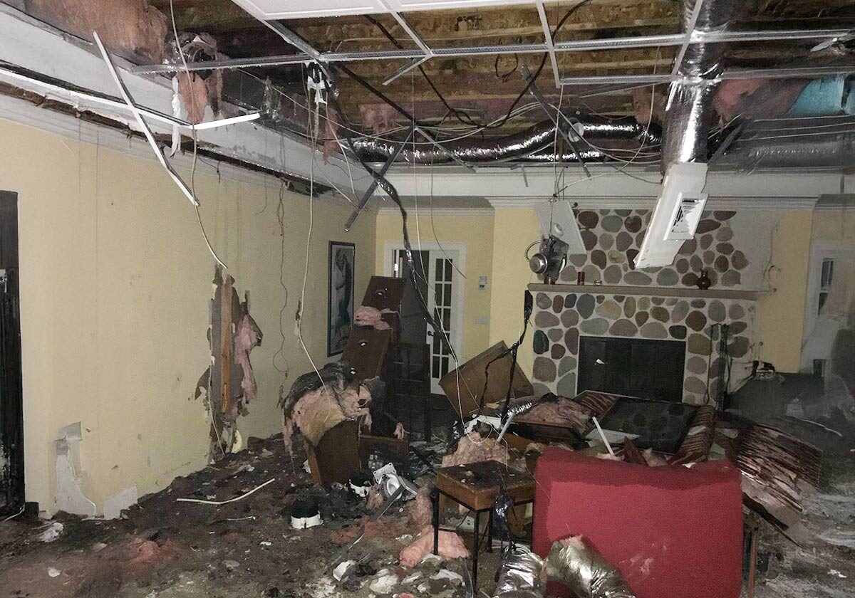 A Living Room Showing Significant Damage With A Collapsed Ceiling, Exposed Beams, And Insulation Hanging Down. Debris Covers The Floor, And Furniture Is Overturned. A Stone Fireplace Is Visible In The Background.